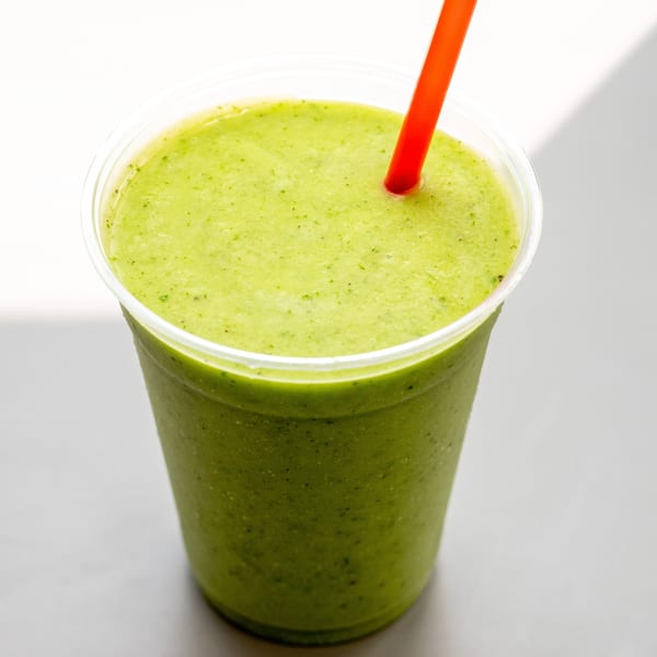 A vibrant green smoothie in a clear plastic cup with an orange straw, on a white surface.